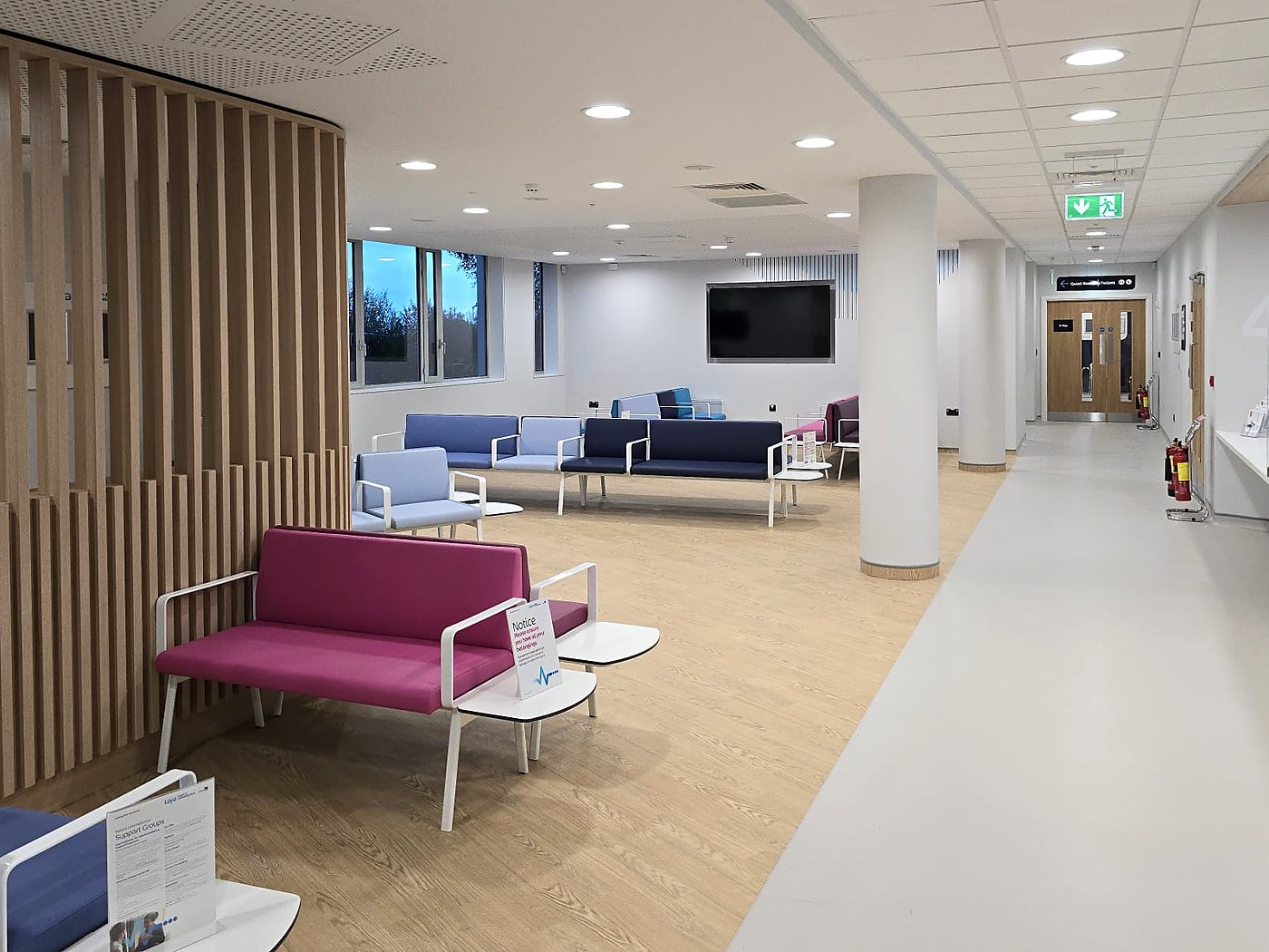 The waiting area of a Laya Health & Wellbeing Clinic: a timber slatted screen, blue and pink seating beside a window, and a bright corridor leading towards reception.